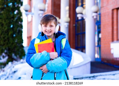 Schoolboy Holds Notebooks In His Hands. The Boy Stands Against The Backdrop Of The School In Winter. Child In Winter Clothesholds Notebooks. Goes To School