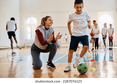 Schoolboy leading soccer ball among cones while sports teacher is supporting him during physical education class. Focus is on teacher.  - Powered by Shutterstock