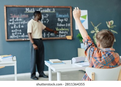 Schoolboy raising hand while Black male teacher explaining math lesson at chalkboard in classroom, showing active learning and participation. Ideal for education marketing and school promotion