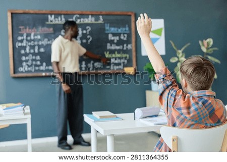 Schoolboy raising hand while Black male teacher explaining math lesson at chalkboard in classroom, showing active learning and participation. Ideal for education marketing and school promotion