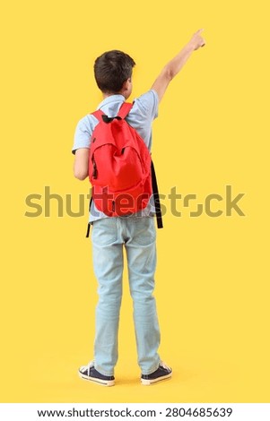Schoolboy with red backpack pointing somewhere on orange background, back view
