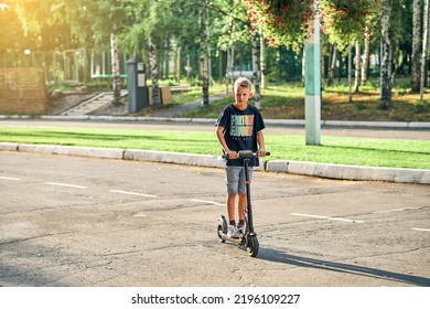 Schoolboy Rides Electric Scooter On Road In Empty Summer Park. Child In Black T-shirt And Denim Shorts Rides On Scooter Along Marked Asphalt Road