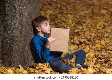 Schoolboy Sitting Near Tree In Fallen Yellow Leaves And Holding Book. Copy Space. Mockup.