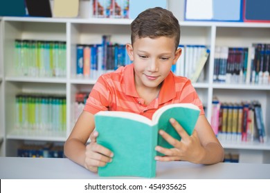 Schoolboy sitting on table and reading book in library at school