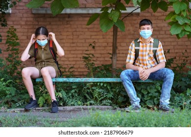 Schoolchildren A Boy And A Teenage Girl Are Sitting On A Bench Outside The School Building, They Use A Protective Breathing Mask Against Covid Or Other Respiratory Infection