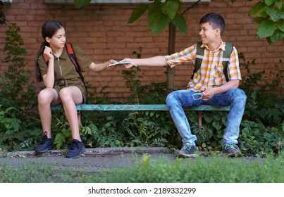 Schoolchildren A Boy And A Teenage Girl Are Sitting On A Bench Outside The School Building, The Boy Gives A Protective Breathing Mask Against Covid Or Other Respiratory Infection