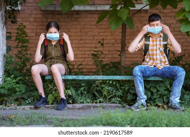 Schoolchildren A Boy And A Teenage Girl Are Sitting On A Bench Outside The School Building, They Use A Protective Breathing Mask Against Covid Or Other Respiratory Infection