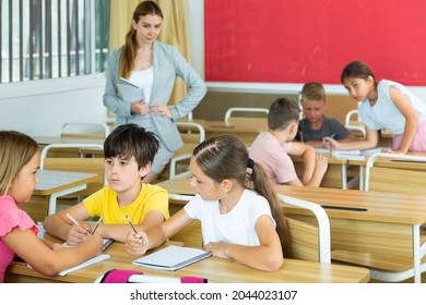Schoolchildren performing group tasks in classroom. Teacher standing beside and observing. - Shutterstock ID 2044023107