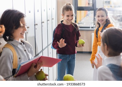 Schoolgirl With Apple And Backpack Waving Hand To Friends On Blurred Foreground In School Hall