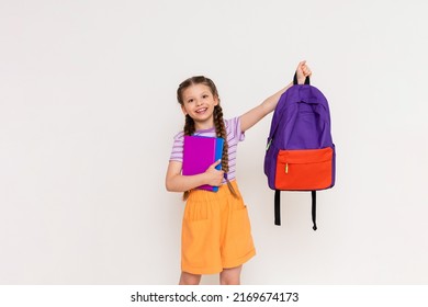 A schoolgirl with books in her hands lifts up a school backpack on a white isolated background. Preparation for the academic year. - Powered by Shutterstock