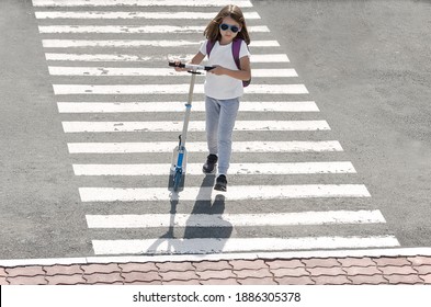 Schoolgirl carrying scooter and crossing road on way to school. Zebra traffic walk way in the city. Pedestrian passing a crosswalk. Stylish child walking with backpack. Safety concept for road users