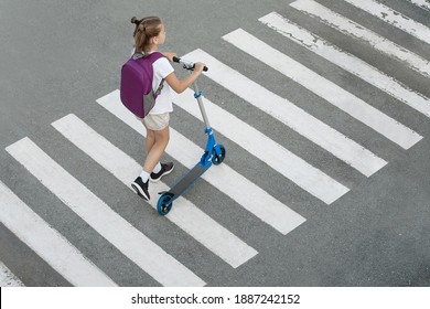 Schoolgirl carrying scooter and crossing road on way to school. Zebra traffic walk way in the city. Pedestrian passing a crosswalk. Stylish child walking with backpack. Safety concept for road users - Powered by Shutterstock