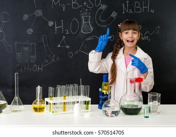 schoolgirl in chemistry lab in white gown pointing at formula on blackboard near table with reagents