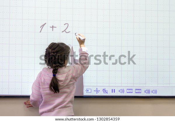 Schoolgirl in the classroom writes on\
a whiteboard with a touch screen. solves an example in\
math