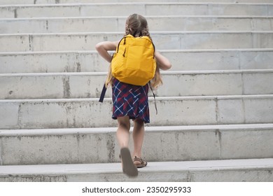 Schoolgirl in colorful dress with bright yellow backpack goes to school little girl with long braids climbs concrete steps on way to class backside view