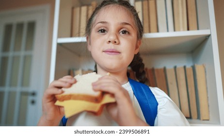 schoolgirl eats a sandwich at school during recess with backpack and bookcase. child lunch education concept. child in classroom having lifestyle lunch snack with bread and cheese sandwich