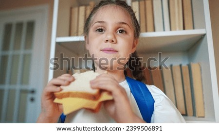 schoolgirl eats a sandwich at school during recess with backpack and bookcase. child lunch education concept. child in classroom having lifestyle lunch snack with bread and cheese sandwich