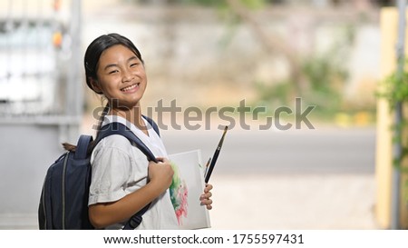 A schoolgirl is holding painting equipment and carrying a school bag while standing and waiting for a school bus.