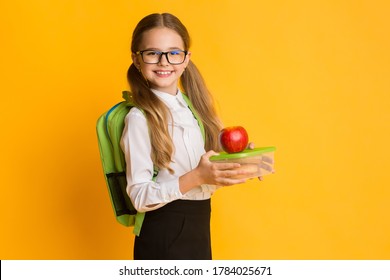 Schoolgirl Holding School Lunch Box And Red Apple Posing On Yellow Studio Background. Free Space For Text