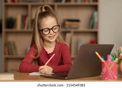 Schoolgirl Learning Online Using Laptop And Taking Notes Sitting At Desk At Home. Girl Writing Looking At Computer Wearing Glasses Having Remote Class Near Bookshelf In Library. E-Learning Concept