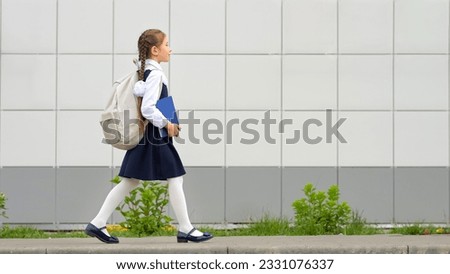 Schoolgirl with long hair carrying white backpack and blue book goes to modern school. Elementary student with braids starts new day at school, side view