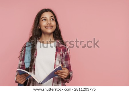 Schoolgirl with open notebook and backpack looking at copy space for advertising on pink studio background, education, learning and school life concept