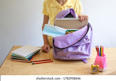 A Schoolgirl Stands Next To A Table With School Supplies And An Open Backpack With A Medical Protective Mask Inside