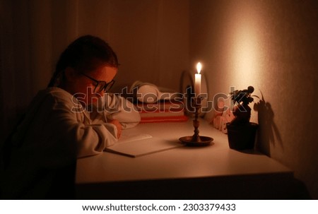 Schoolgirl uses candlelight to writes and reads in darkness without electric lights at home. Teen with poor eyesight wears glasses to studies and to do school homework during blackout.Vision problems.