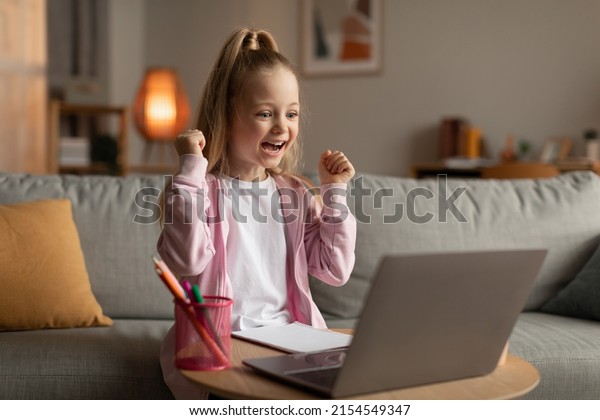Schoolgirl Using Laptop Shaking Fists Celebrating\
Educational Success, Gesturing Yes Sitting Near Computer At Home.\
E-Learning Achievements. Distance School And Internet Education.\
Side View