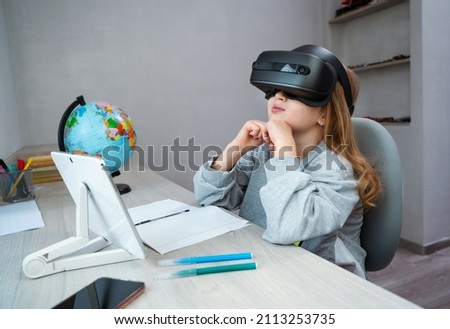 Schoolgirl using VR headset simulators for studying. Child is wearing virtual reality glasses sitting at desk with tablet and watching on top. Future learning concept