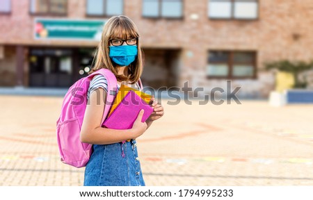 Schoolgirl wearing face mask during epidemic. Back to school concept. Cute girl outside at school having good time. Safety mask to coronavirus prevention. Kid with backpack going to school. Education.