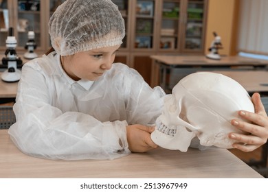 A schoolgirl in a white coat studies an anatomical model of a human skull in a school classroom. Anatomy lesson. High quality photo - Powered by Shutterstock