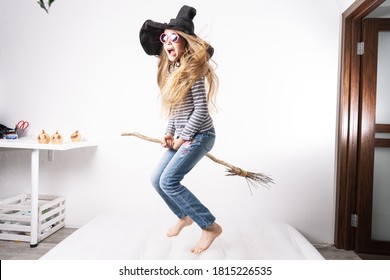 Schoolgirl in a witch hat jumps on a mattress and flies on a broomstick. Children's lifestyle portrait.