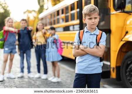 Schoolkids bullying their upset classmate while standing near school bus, vicious children pointing at boy and laughing, upset male child feeling lonely and depressed, selective focus