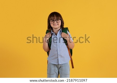 Schooltime concept. Studio portrait of little clever boy preschooler wearing eyeglasses smiling to camera with backpack, yellow background