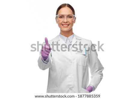 science and profession concept - happy smiling female scientist in goggles and gloves with nametag on lab coat showing thumbs up