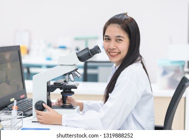 Scientist Asian woman smiling and sitting with microscope in laboratory.Researcher using microscope in medical laboratory. Medical healthcare technology and pharmaceutical research concept.