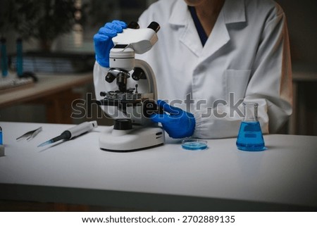 Scientist in blue gloves operating a microscope on a white lab bench with various scientific equipment, performing chemical research
