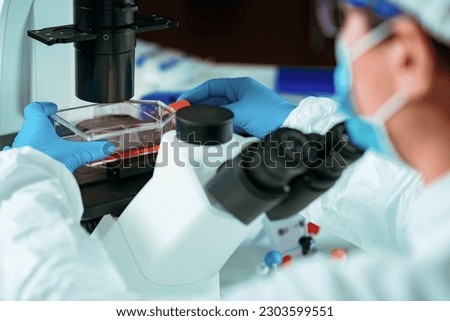 A scientist carefully places a flask filled with cells onto a microscope plate. The lab is illuminated with bright white light
