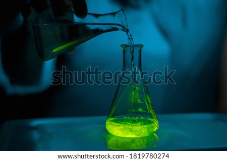 A scientist experimenting with a green fluorescent solution in a glass conical flask in dark biomedical laboratory for health care development
