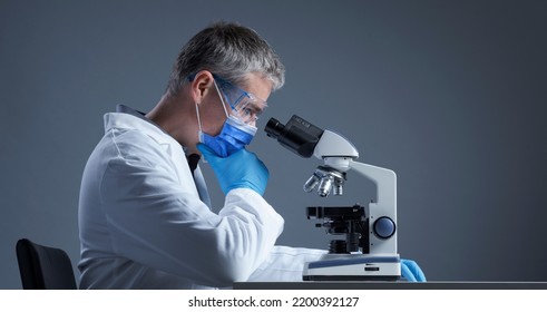 Scientist with face mask examining a sample under the microscope and thinking, scientific research concept, blank copy space - Shutterstock ID 2200392127