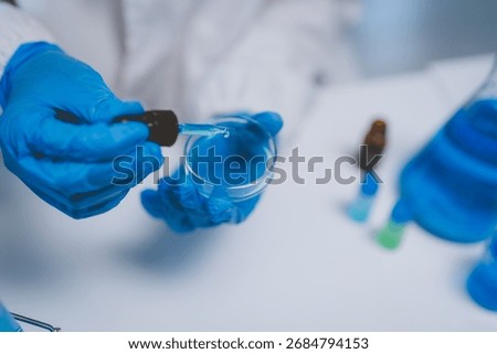 Scientist hands in blue gloves adding a drop of blue liquid to a petri dish, conducting a chemical experiment in a lab