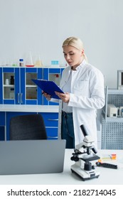 Scientist Holding Clipboard Near Laptop And Microscope In Lab