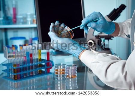 A scientist holding a petri dish in the lab with a monitor and microscope in background.
