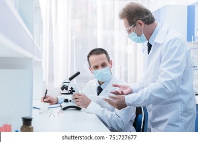 Scientist In Lab Coat And Sterile Mask Doing Microscope Analysis And Talking To A Colleague