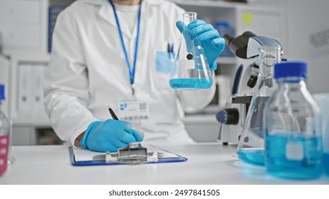 A scientist in a lab writes observations while analyzing a blue liquid in a flask, exemplifying research in a clinic. - Powered by Shutterstock