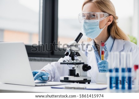 scientist in medical mask and goggles holding test tube with covid lettering and using laptop