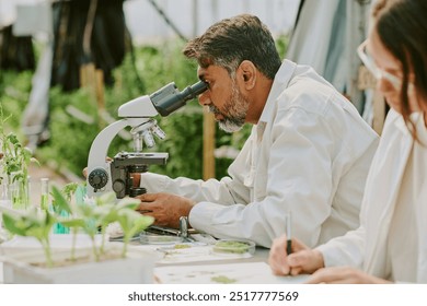 Scientist observing plant samples through a laboratory microscope in greenhouse environment. Colleague working alongside, preparing scientific materials on table - Powered by Shutterstock