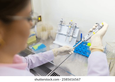 Scientist Pipetting Sample into Test Tube in Laboratory Hood. Multiple pipettes are mounted on a stand, and petri dishes are stacked in the background, highlighting a sterile laboratory environment.