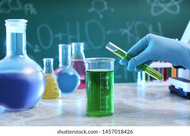 Scientist Pouring Liquid Into Beaker At Table Against Chalkboard, Closeup. Chemistry Glassware